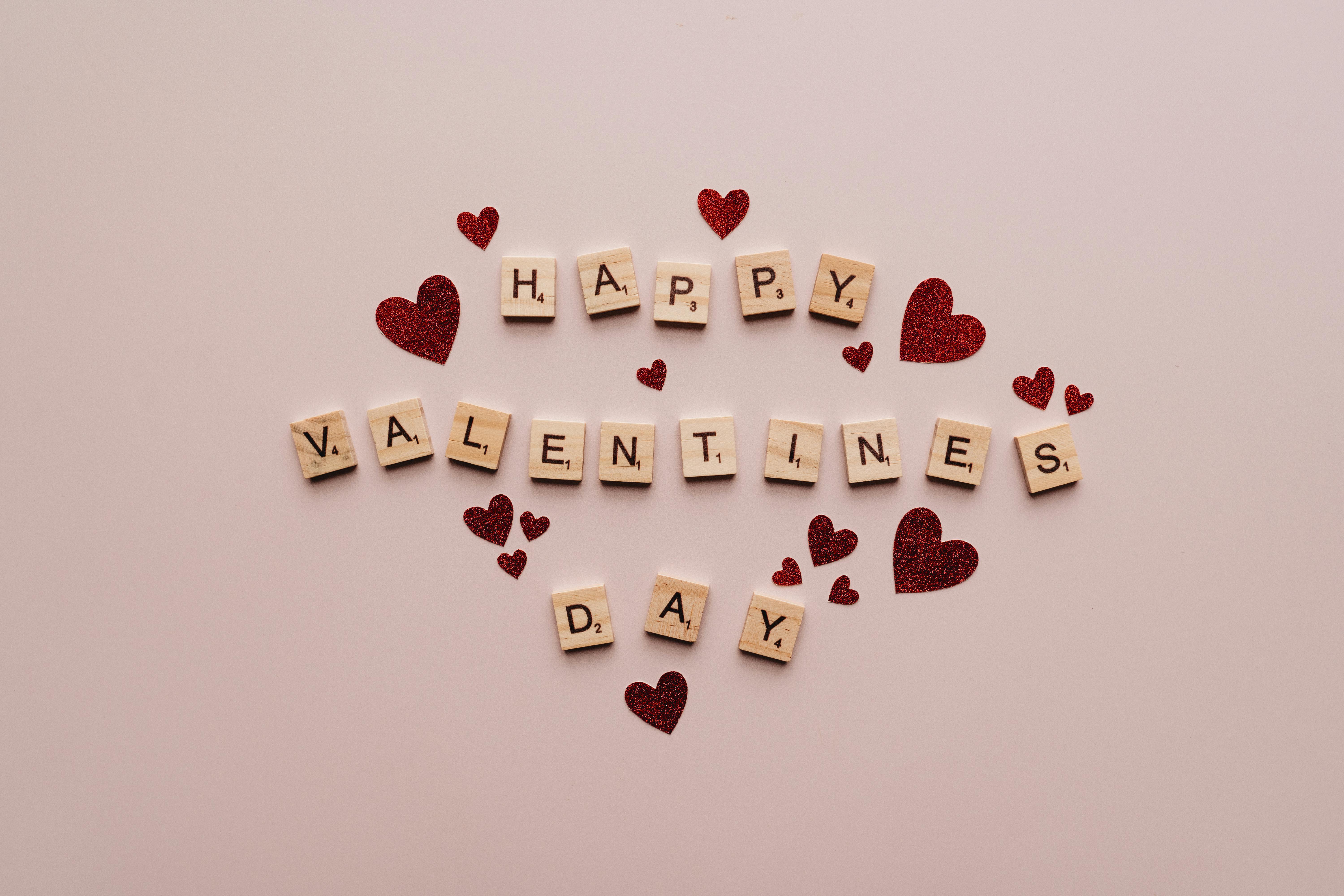 A festive Valentine's Day greeting with Scrabble tiles and red glitter hearts on a soft background.