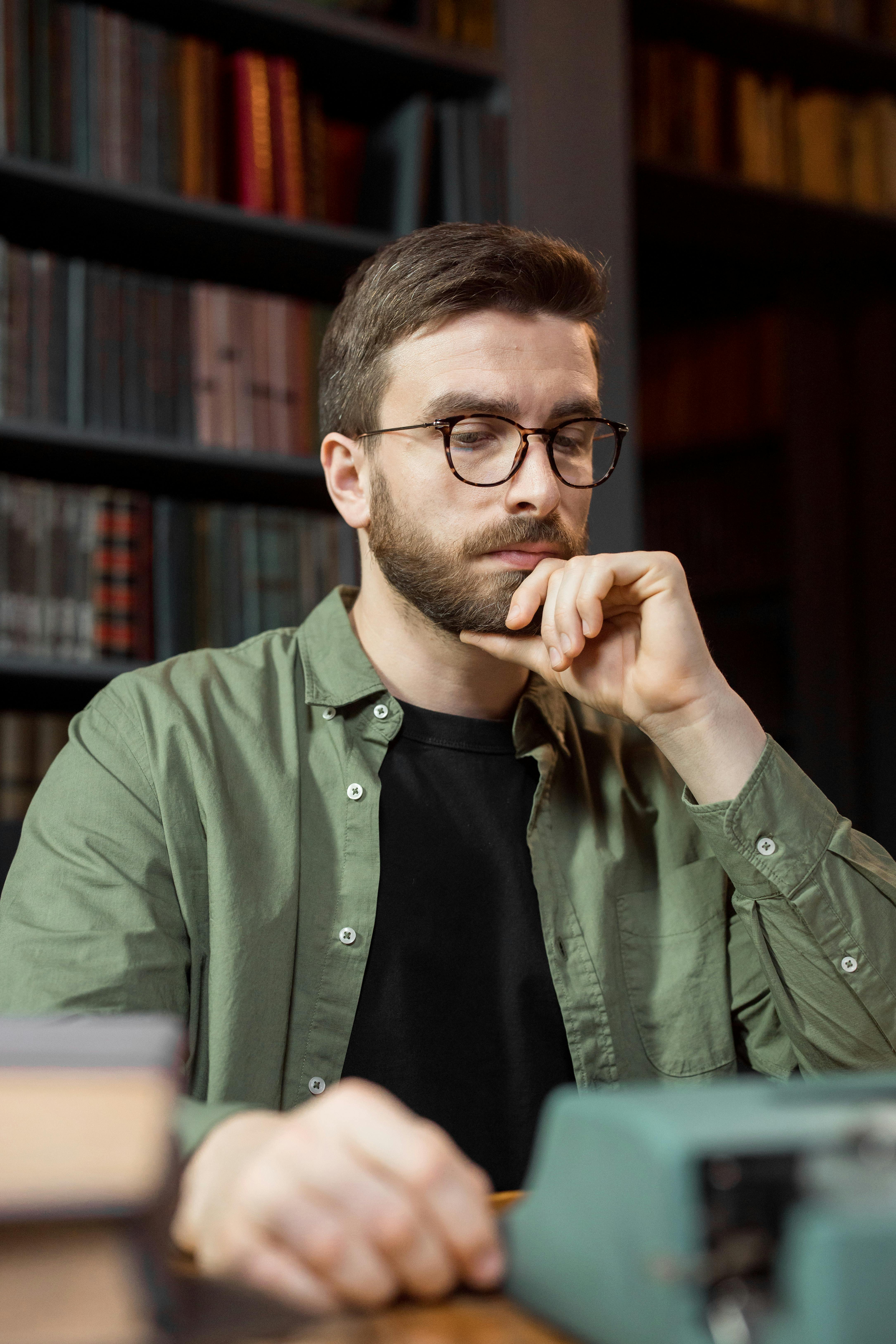 Man in glasses thinking deeply while using typewriter in library setting.